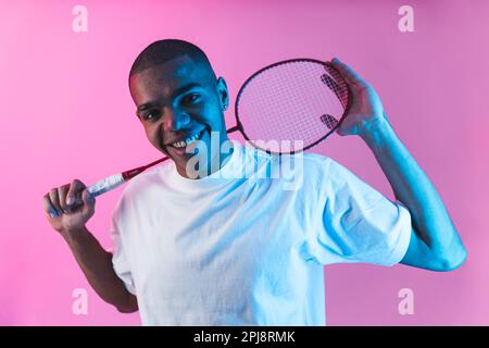 Homme avec raquette de badminton tourné en studio. Jeune homme souriant, charmant, tenant l'équipement de badminton derrière son cou et regardant l'appareil photo sur fond rose. Photo de haute qualité Banque D'Images