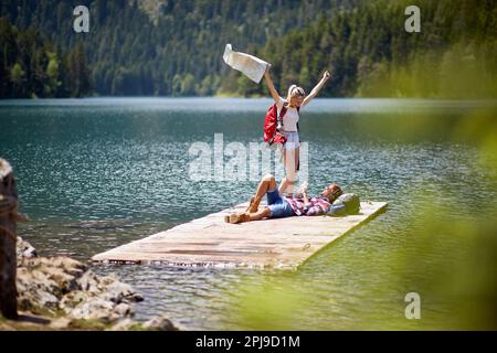 Backpackers sur une jetée en bois au bord du lac. Femme excitée avec la carte, homme couché à l'écoute de la musique et souriant. Concept de style de vie, de convivialité, de nature Banque D'Images