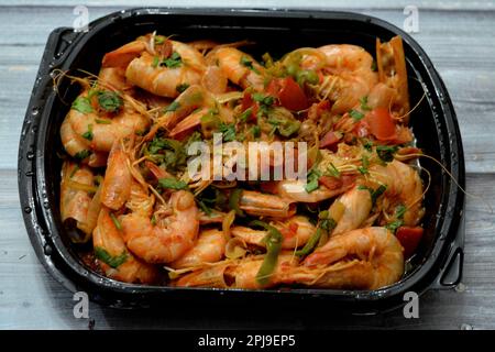 Crevettes cuites mélange d'ail, de citron, de persil, de poivre, d'oignon et de tomate servi dans une assiette noire jetable isolée sur fond de bois, foyer sélectif Banque D'Images