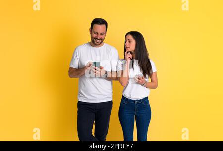 Curieuse jeune femme adulte avec la main sur le menton pélécher dans le téléphone mobile de l'ami et essayant de lire ses messages sur fond jaune Banque D'Images