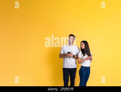 Une petite amie adulte jalouse qui se tourne sur le smartphone de l'ami et regarde loin tout en se tenant sur fond jaune Banque D'Images