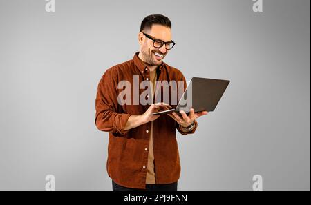 Jeune homme d'affaires souriant et concentré, habillé d'une chemise, travaillant sur un ordinateur portable tout en restant en confiance sur un arrière-plan gris isolé Banque D'Images