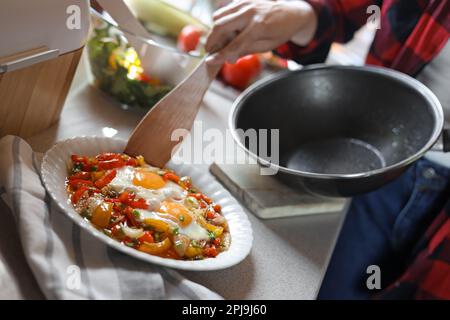 Femme mettant des œufs fraîchement frits avec des légumes sur l'assiette dans la cuisine, gros plan Banque D'Images