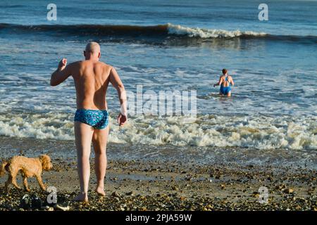 Homme dans les trunks courant dans la mer avec chien sur Avon Beach Christchurch, Royaume-Uni en hiver Banque D'Images