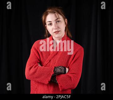 Jeune femme caucasienne debout avec un petit sourire, bras pliés, portant un gilet rouge sur une robe noire devant un fond noir Banque D'Images