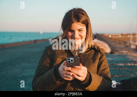 Une femme caucasienne confiante et de grande taille marche seule sur une jetée en bois au bord de la mer, souriant alors qu'elle vérifie son smartphone. La plage sereine et l'arrière-pays de l'océan Banque D'Images