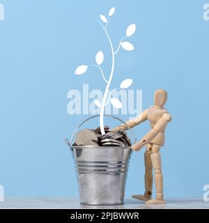 Un mannequin en bois et un seau miniature avec des pièces de monnaie en métal et un arbre peint avec des feuilles sur fond bleu. Concept de croissance des revenus et des bénéfices Banque D'Images