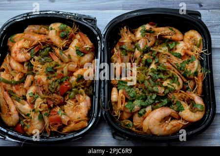 Crevettes cuites mélange d'ail, de citron, de persil, de poivre, d'oignon et de tomate servi dans une assiette noire jetable isolée sur fond de bois, foyer sélectif Banque D'Images