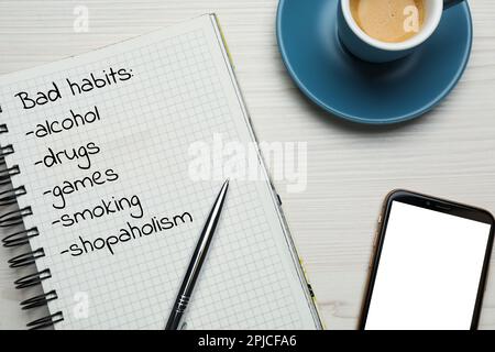 Carnet avec liste de mauvaises habitudes, tasse de café et smartphone sur table en bois blanc, plat Banque D'Images