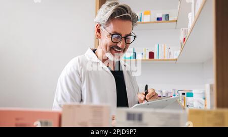 Un pharmacien de sexe masculin écrit sur une planchette à pince tout en distribuant des ordonnances dans un pharmacien. Travailleur de la santé senior travaillant dans une pharmacie. Banque D'Images