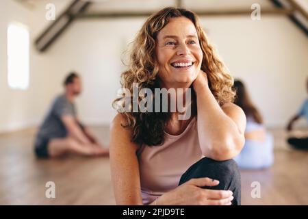 Femme aînée souriante assise dans un studio de yoga avec un groupe de personnes s'exerçant en arrière-plan. Bonne femme mûre ayant une séance d'entraînement avec lui Banque D'Images