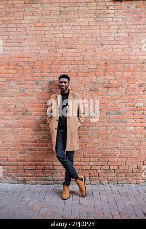 Homme afro-américain élégant et confiant regardant la caméra tout en posant à l'extérieur contre un mur de briques. Banque D'Images