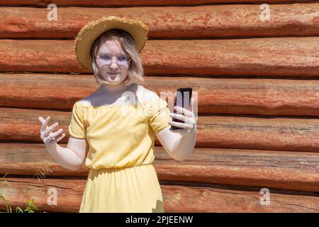 Asiatique adolescente en robe jaune, chapeau et lunettes de soleil a appel vidéo, chat en ligne contre le mur de la maison de village en bois, espace de copie. Heure d'été. Hol Banque D'Images