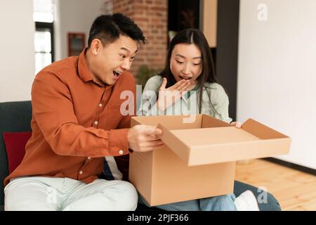 Surpris homme asiatique et femme regardant à l'intérieur de boîte en carton, satisfait avec l'article commandé, intérieur de salon Banque D'Images