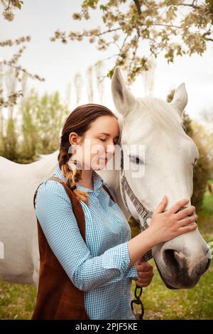 Une belle jeune femme pose à côté d'un cheval blanc. Cowboy, ranch, village. Amour amitié préoccupation. Animaux de compagnie. À l'extérieur de la porte, bois d'herbe verte. sunlig Banque D'Images