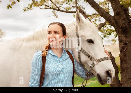 Une belle jeune femme pose à côté d'un cheval blanc. Cowboy, ranch, village. Amour amitié préoccupation. Animaux de compagnie. À l'extérieur de la porte, bois d'herbe verte. sunlig Banque D'Images