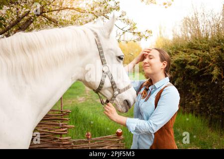 Une belle jeune femme pose à côté d'un cheval blanc. Cowboy, ranch, village. Amour amitié préoccupation. Animaux de compagnie. À l'extérieur de la porte, bois d'herbe verte. sunlig Banque D'Images