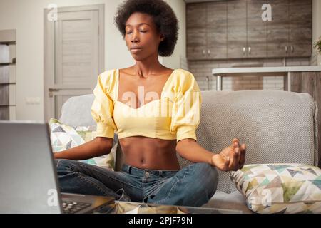 Cours de méditation en ligne, calme sur Internet, calmant, communication. Une femme noire afro-américaine à la maison avec un ordinateur portable. Banque D'Images