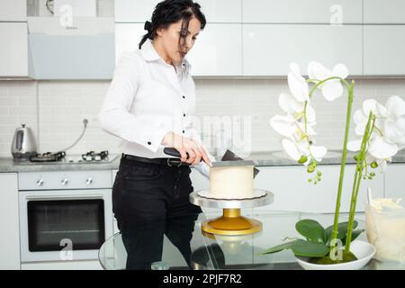 Le confiseur évisse le gâteau, le couvre avec du chocolat blanc. Production à domicile, affaires, entrepreneur, passe-temps. Pâtisseries et bonbons délicieux. kit blanc Banque D'Images