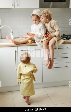 Une fille et un garçon dans des peignoirs sont assis dans la cuisine et tiennent des oranges confites dans leurs mains, le plus jeune enfant les regarde. Banque D'Images