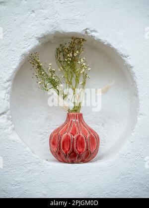 Fleurs sauvages dans un vase rouge, logée dans un hublot contre un mur blanc, dans un lieu de mariage Banque D'Images