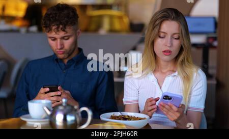 Un jeune homme et une jeune femme mécontents assis dans un café et utilisant un smartphone. Couple tapant sur le téléphone mobile, ne parlant pas les uns aux autres . Problème de relation Banque D'Images