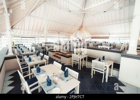 Photo grand angle d'un restaurant paisible des Maldives divisé en deux niveaux, avec des meubles blancs, des serviettes et des coussins bleus, un sol noir, et un ouvert Banque D'Images