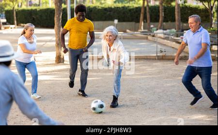 Joyeux amis multinationaux jouant au football sur un terrain de jeu sablonneux lors d'une chaude soirée de printemps Banque D'Images