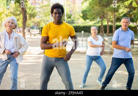Un entraîneur afro-américain positif aidant les personnes âgées à étirer leurs muscles avant de danser à l'extérieur dans le parc Banque D'Images