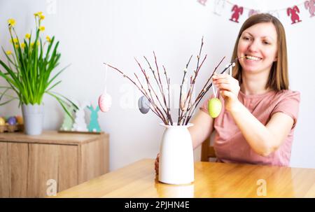 Femme souriante décorant un bouquet de brunches de saule avec des œufs de Pâques colorés à la maison. Banque D'Images