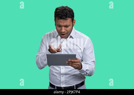 Portrait d'un Indien brun en colère contre quelqu'un en ligne. Homme d'affaires avec tablette pc sur fond vert. Banque D'Images