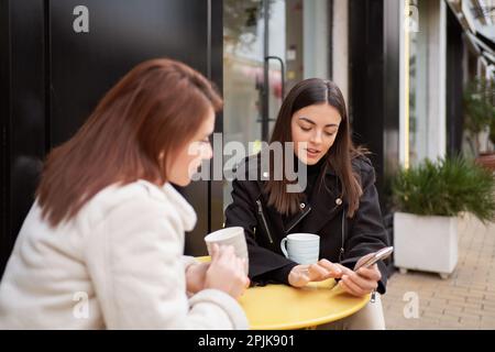 Deux femmes utilisant un téléphone portable tout en buvant du café au café. Technologie et amitié. Banque D'Images