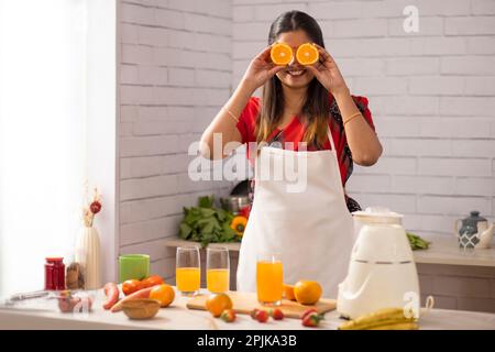 La femme heureuse couvre les yeux avec des tranches d'orange tout en faisant du jus d'orange dans la cuisine Banque D'Images