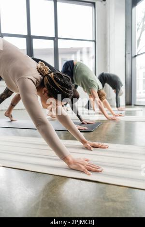 Femme afro-américaine pratiquant le chien face à la baisse pose sur le tapis dans la classe de yoga, image de stock Banque D'Images