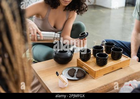 vue rognée d'une femme qui verse de l'eau chaude à partir de thermos tout en préparant du thé pu-erh près des gens dans un studio de yoga, image de stock Banque D'Images