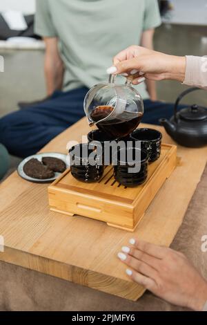 vue partielle de la femme qui verse le puer filtre de la verseuse en verre dans les tasses à thé traditionnelles, image de stock Banque D'Images