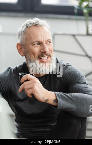 Positif homme d'âge moyen avec tatouages tenant une tasse chinoise avec du thé de puer dans le studio de yoga, image de stock Banque D'Images