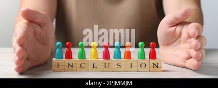 Femme protégeant les pions colorés et les cubes en bois avec le mot inclusion à la table blanche en bois, gros plan. Bannière Banque D'Images