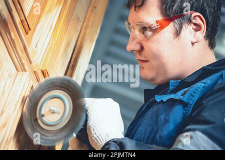 L'homme traite la surface en bois. Le travailleur dans les lunettes de sécurité grince la porte en bois avec une brosse métallique. Réparation professionnelle de meubles et de produits en bois. Banque D'Images