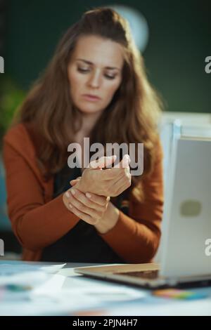 Gros plan sur une femme d'âge moyen propriétaire d'une petite entreprise avec un ordinateur portable et une douleur au poignet dans le bureau vert. Banque D'Images