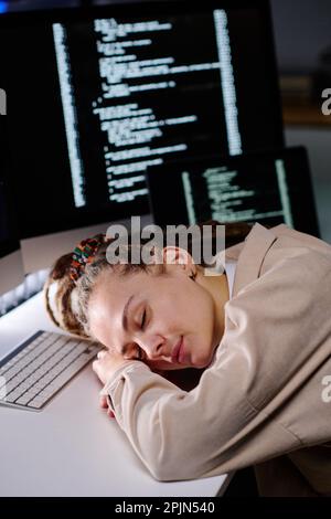 Jeune femme fatiguée avec des dreadlocks gardant la tête sur le bureau par le clavier tout en dormant dans le bureau sombre contre des écrans d'ordinateur avec des données codées Banque D'Images