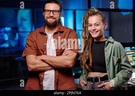 Des jeunes ingénieurs INFORMATIQUES, hommes et femmes, se tenant devant l'appareil photo et vous regardant avec le sourire tout en travaillant tard dans le bureau sombre Banque D'Images