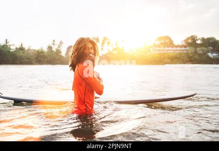 Portrait d'un adolescent noir à poils longs avec planche de surf prête pour le surf avec rétroéclairage au coucher du soleil. Il marche dans les vagues de l'océan Indien. Sports nautiques extrêmes Banque D'Images