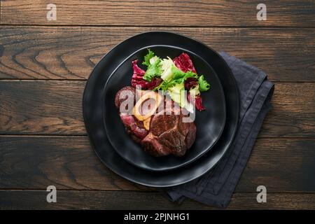 Steaks Osso Buco. Bœuf grillé Osso Buco ou steak de viande de portreuse avec garniture de salade et de frites sur un panneau de marbre noir sur du vieux bois Banque D'Images