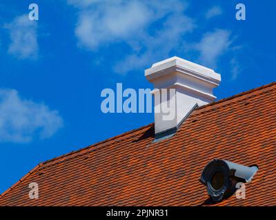 cheminée en stuc blanc. toit incliné avec tuiles de toit en argile brun rouge. plaque de zink ovale recouverte de dormeur et de solin. ciel bleu et nuages blancs. Banque D'Images