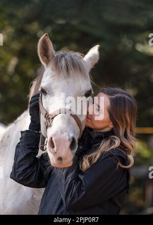 Jeune femme debout à côté des yeux blancs de cheval arabe fermé comme si elle s'embrasse ou sentait, brouillé arbre arrière-plan, gros plan Banque D'Images