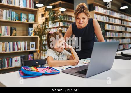 Petite fille étudiante faisant ses devoirs en utilisant un ordinateur portable dans un club après l'école primaire. Professeur aidant à résoudre et à terminer la tâche. Retour à l'école. Banque D'Images