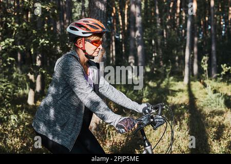 Femme active à vélo sur la route forestière. Une femme qui fait du vélo tout-terrain le jour des vacances d'été. Femme portant un casque de vélo et des gants Banque D'Images