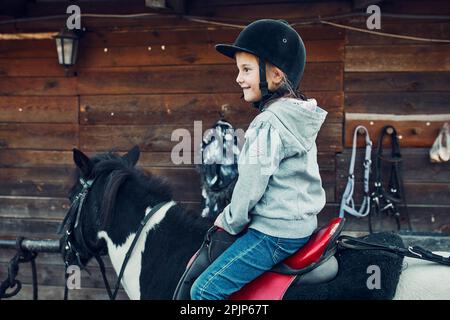 Petite fille souriante apprenant l'équitation. 5-6 ans équestres en casque ayant plaisir à monter un cheval Banque D'Images