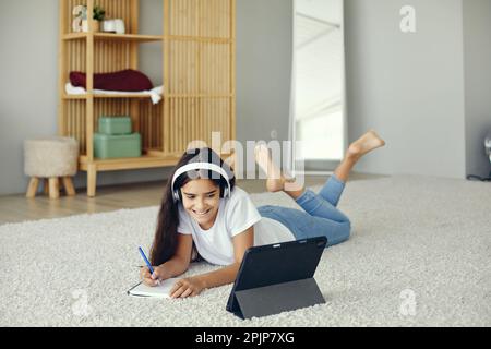 Jolie pré-adolescent 12s fille porter des écouteurs sans fil, couché sur le sol écouter cours audio étudier, à la maison école utiliser la tablette numérique. Utilisation des technologies modernes f Banque D'Images